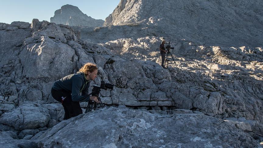 Ein steiles Bergterrain auf dem zwei Menschen mit Kamerastativ etwas außerhalb des Bildes filmen.