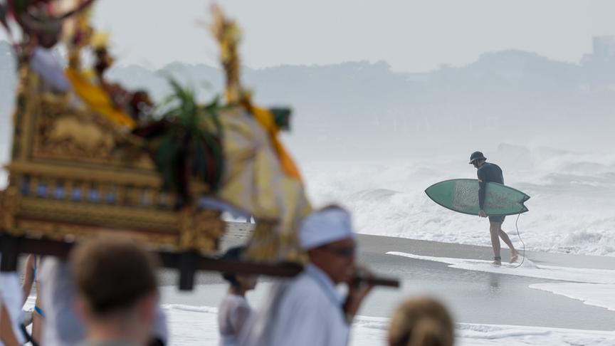 Im Vordergrund eine Gruppe Menschen mit goldenen Zeremoniengegenständen; im Hintergrund ein Surfer, der am Strand entlang geht.