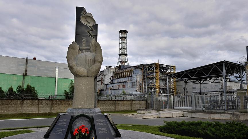 Im Vordergrund: Gedenkstätte mit steinerner Statue, im Hintergrund: Reaktorgebäude umgeben von Metall