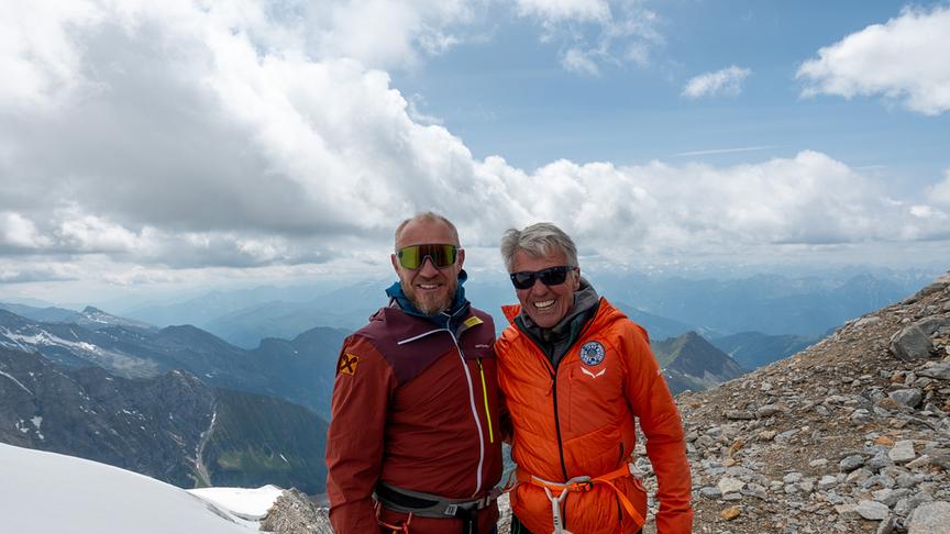 Hermann Maier und Peter Habeler nebeneinander stehend und vor bergigem Hintergrund mit Wolken posierend.