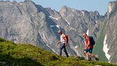 © Foto: ORF/Interspot/Christoph Prochazka Peter Habeler und Hermann Maier beim wandern über grüne Vegetation mit steilem Bergpanorama im Hintergrund.