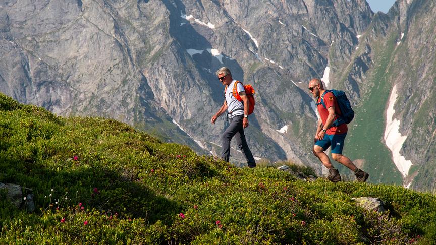 Peter Habeler und Hermann Maier beim wandern über grüne Vegetation mit steilem Bergpanorama im Hintergrund.