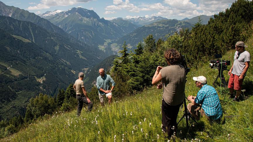 Ein Kamerateam filmt ein Gespräch zwischen Kurt Lechner und Hermann Maier auf einer steil abfallenden Bergwiese. Im Hintergrund ist ein beeindruckendes Bergpanorama unter blau, bewälktem Himmel.