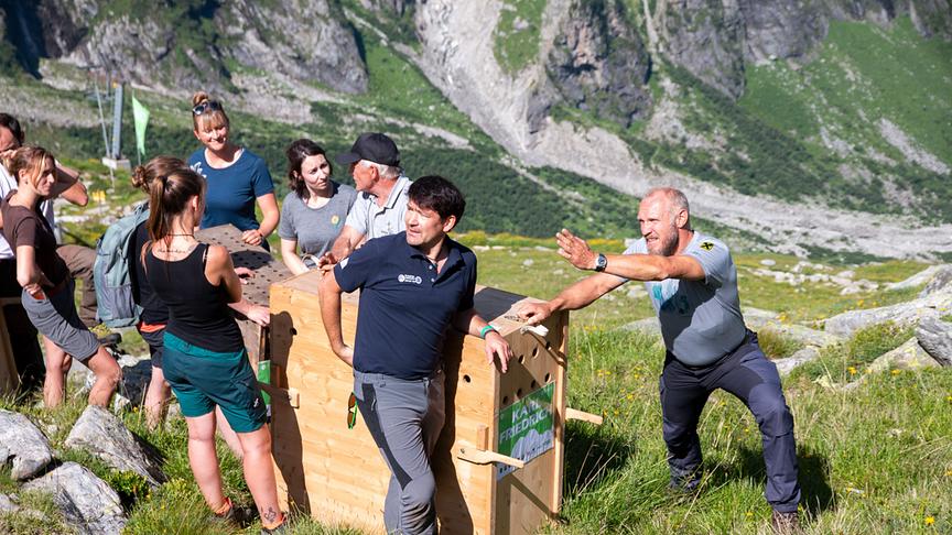 Mehrere Personen schleppen eine hohe Holzbox durch bergiges Gelände.
