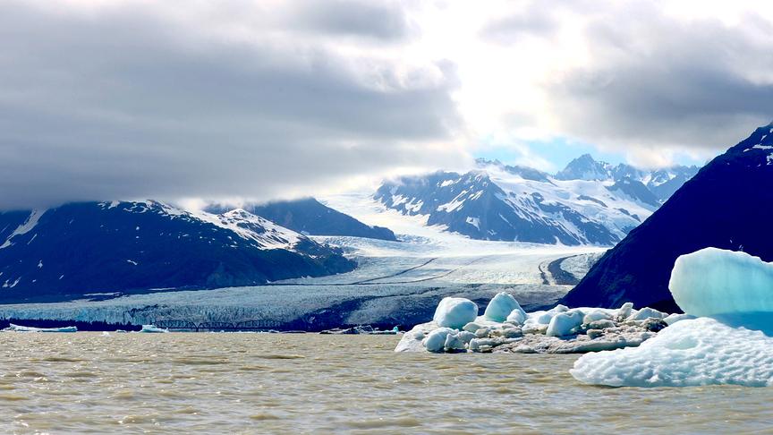 Ein Gletscher zwischen hohen schneebedeckten Bergen, der in einem Gewässer mündet. Die Wolken hängen tief zwischen den Bergen.