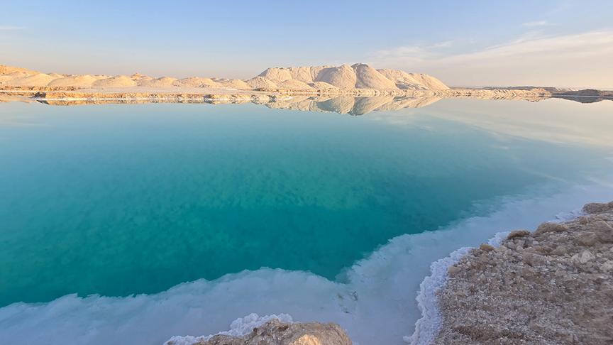 Türkiser See mit Salzablagerungen am Ufer und weißen Salzerhebungen im Hintergrund.
