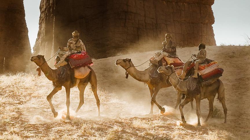 Drei Personen in Camouflage-Kleidung reiten auf Kamelen durch die Wüste.