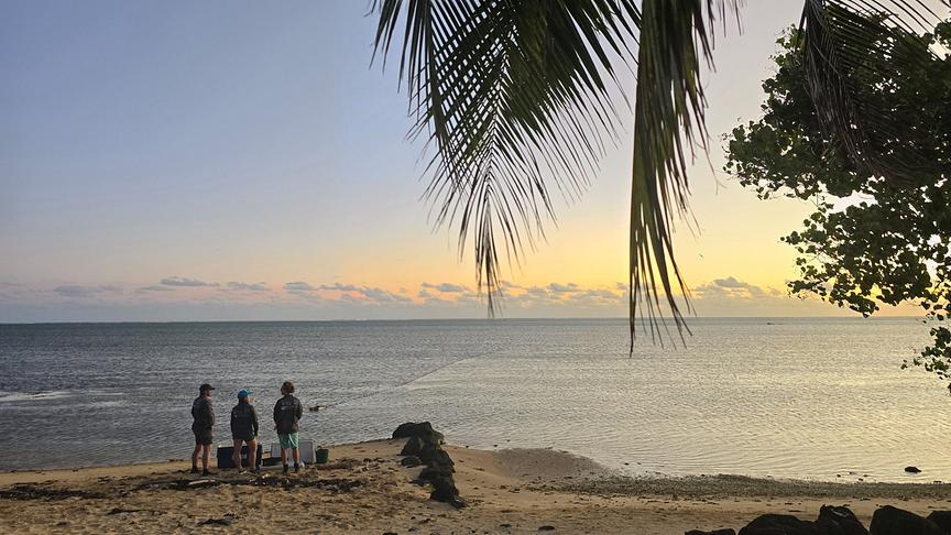 Drei Personen am Strand blicken auf das Meer und den Sonnenuntergang.