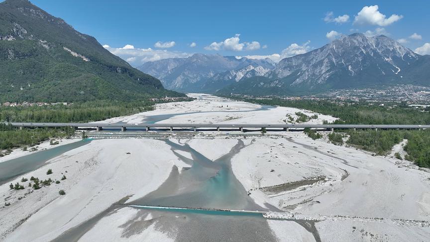 Ein breites steiniges Flussbett, durch den schmale Flussrinnsale laufen, in einem Tal umgeben von hohen Bergen und durchquert von einer Straßenbrücke.