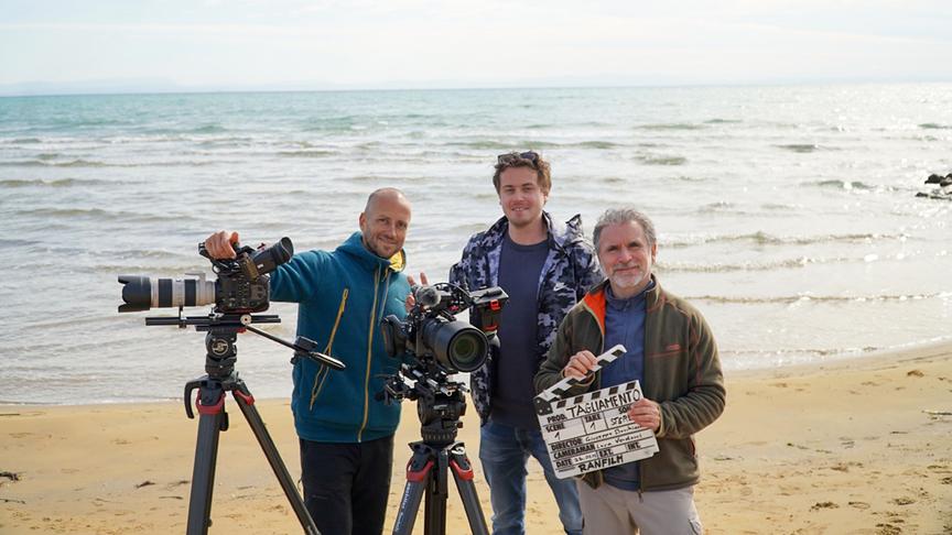 Mehrere Kameramänner mit Equipment und Filmklappe an einem Strand vor dem Meer posierend.