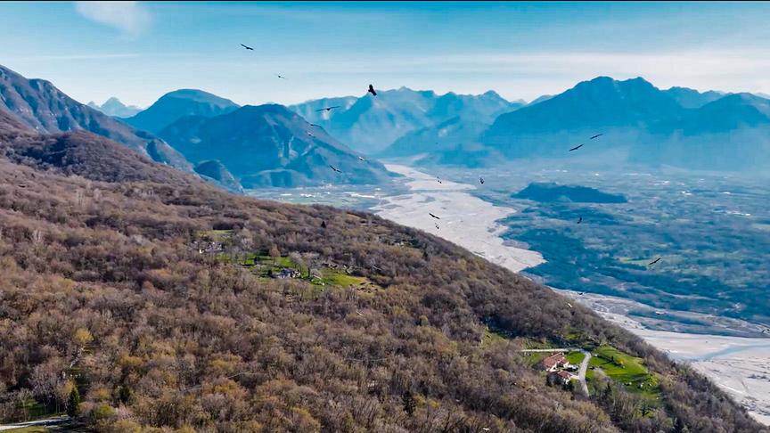 Weite Aufnahme über ein Tal umgeben von Bergen und durchzogen von einem Fluss hinweg. Am Himmel kreisen Geier über die Landschaft.