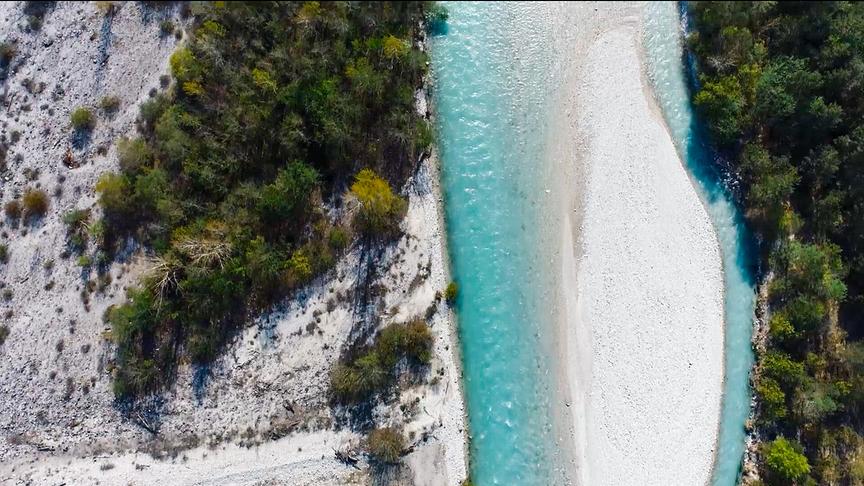 Luftaufnahme von einem türkisen Fluss im steinigen Flussbett. Das Ufer ist sandig und mit grüner Vegetation bewachsen.