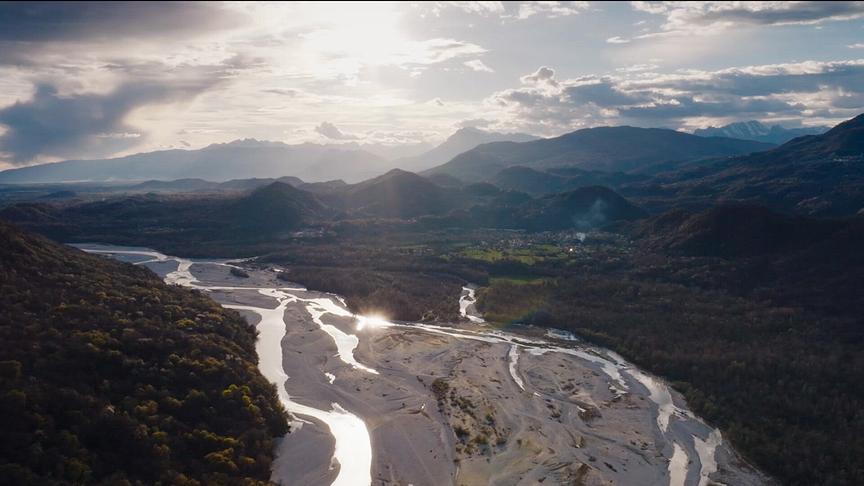 Eine weitläufige Aufnahme von Bergen, Wäldern und einem Fluss mit breitem steinigen Flussbett bei bewölktem Himmel, durch den die Sonne hell scheint.