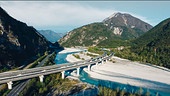 Berglandschaft mit einem Fluss und steinigen Flussbett. Mehere Straßen und eine Brücke durchqueren die Landschaft.