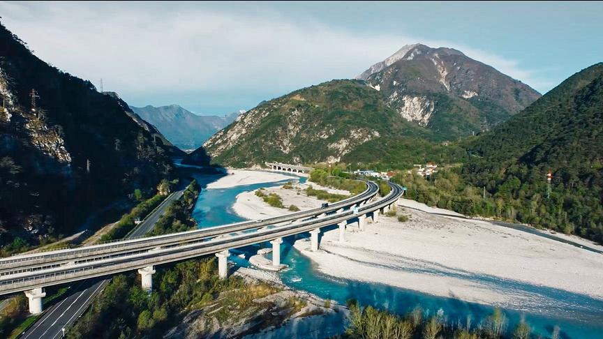 Berglandschaft mit einem Fluss und steinigen Flussbett. Mehere Straßen und eine Brücke durchqueren die Landschaft.