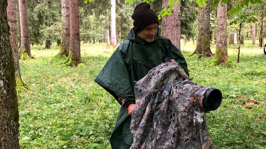Ein Kameramann in Tarn- bzw. Regenkleidung im Wald. Seine Stativkamera ist ebenfalls mit einer Plane in Tarnfarben umhüllt.