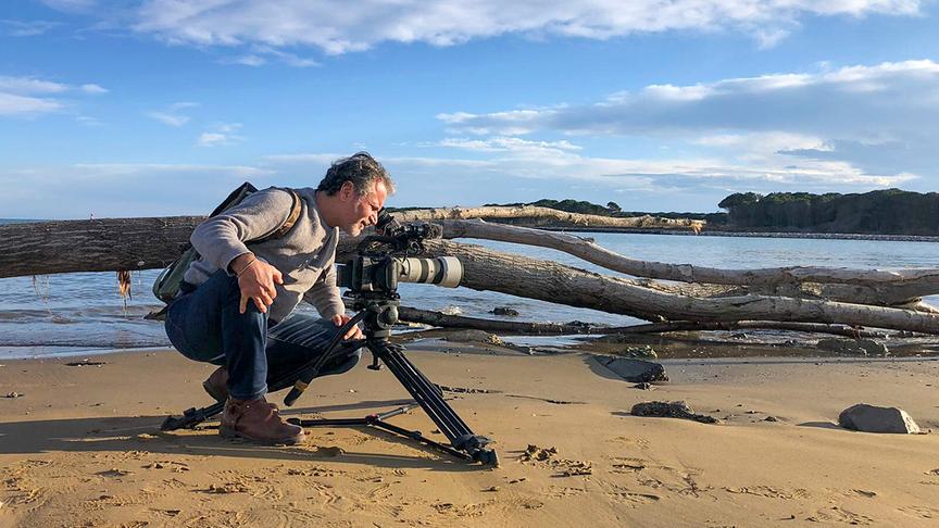 Ein Mann sitzt hinter einer Stativkamera am Boden und filmt an einem Strand. Hinter ihm ist ein umgefallender großer Baum und eine Bucht.