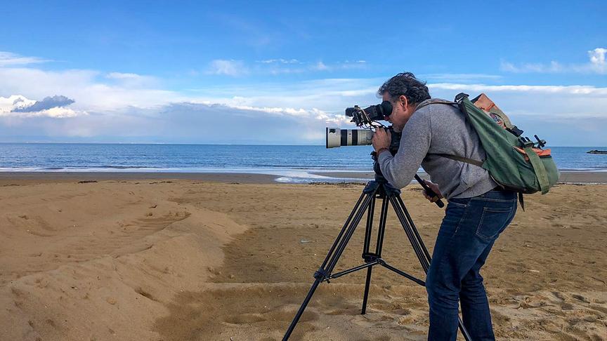 Ein Kameramann film mit einer Stativkamera an einem Strand. Im Hintergrund weites blaues Meer und bewölkter Himmel.