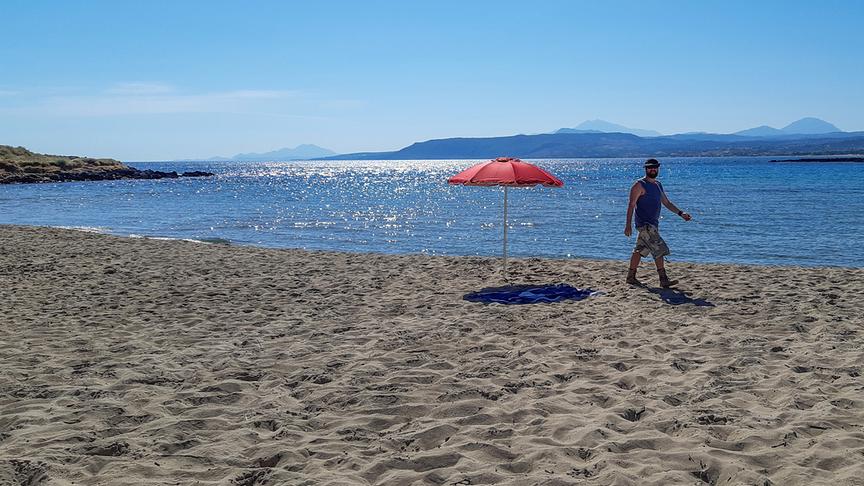 Eine Person geht am Strand entlang, daneben ein einsamer Sonnenschirm, im Hintergrund erheben sich Berge