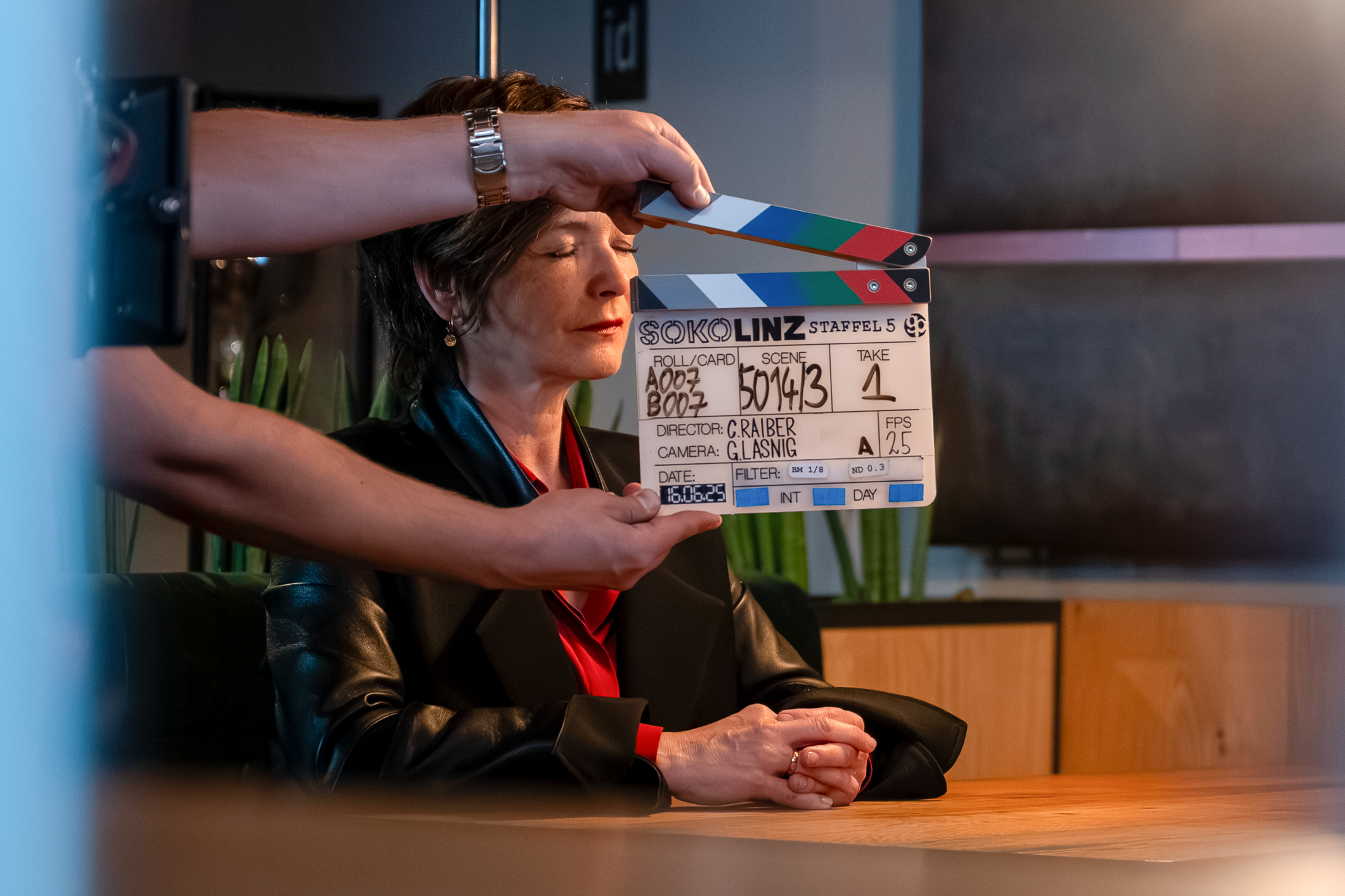 Klappe zu einer Szene der Serie „Soko Linz“ vor Katharina Stemberger, die an einem Tisch sitzt.
