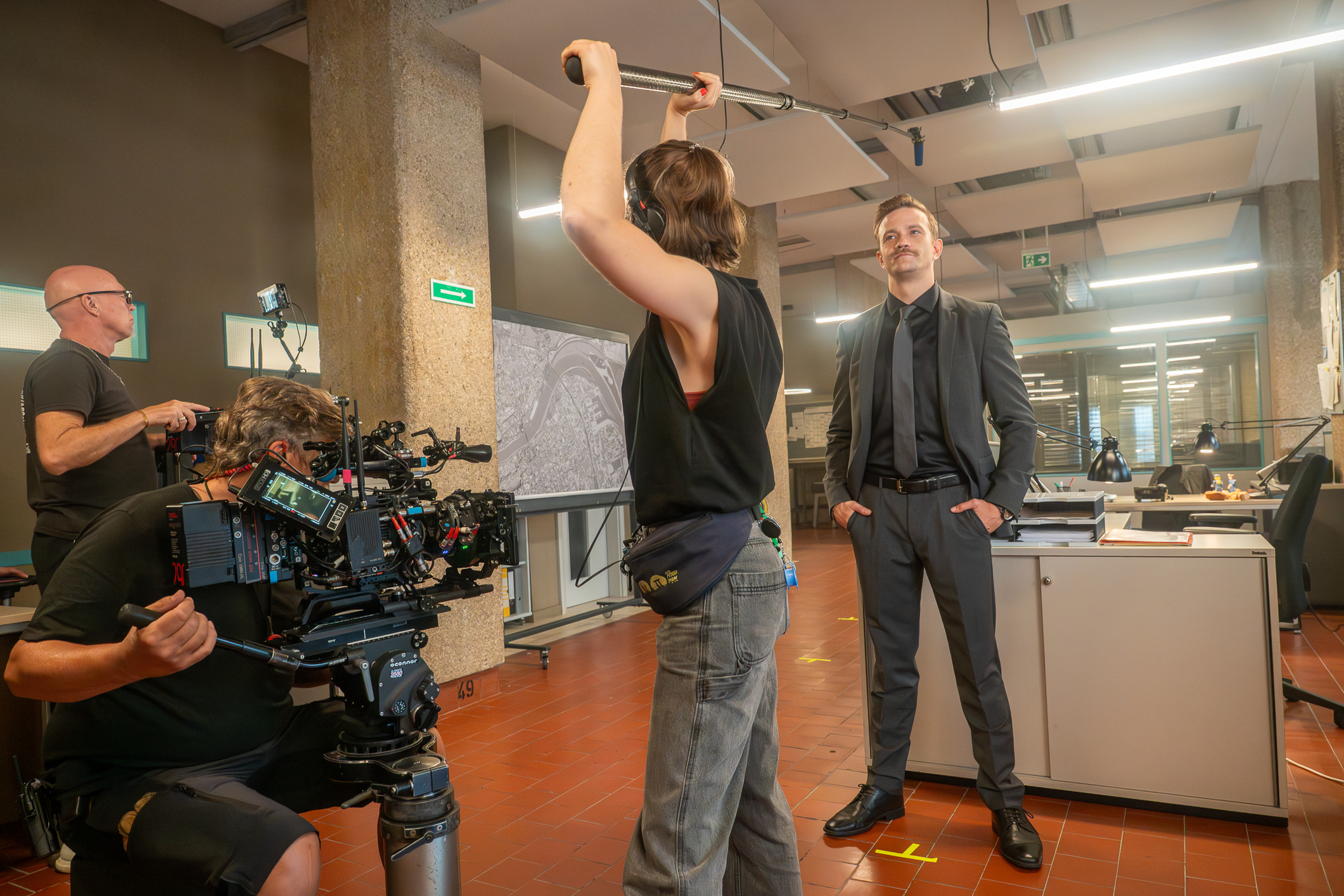 Filmteam dreht eine Szene in einem Büro: Kameraleute und Tonfrau arbeiten, während Michael Steinocher im Anzug vor einem Schreibtisch steht.