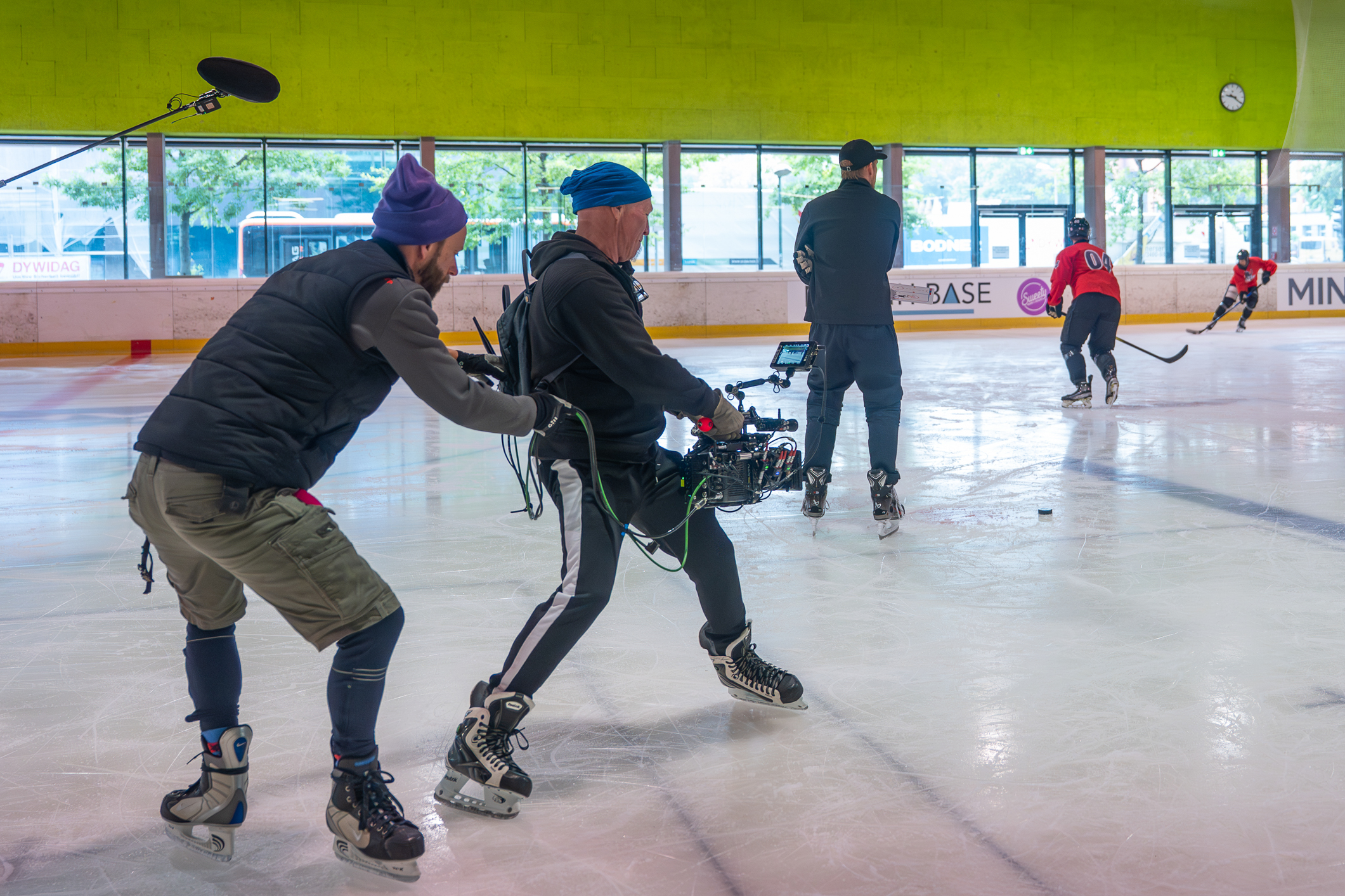 Filmteam auf Eislaufschuhen dreht in einer Eishalle eine Szene mit Eishockeyspielern.