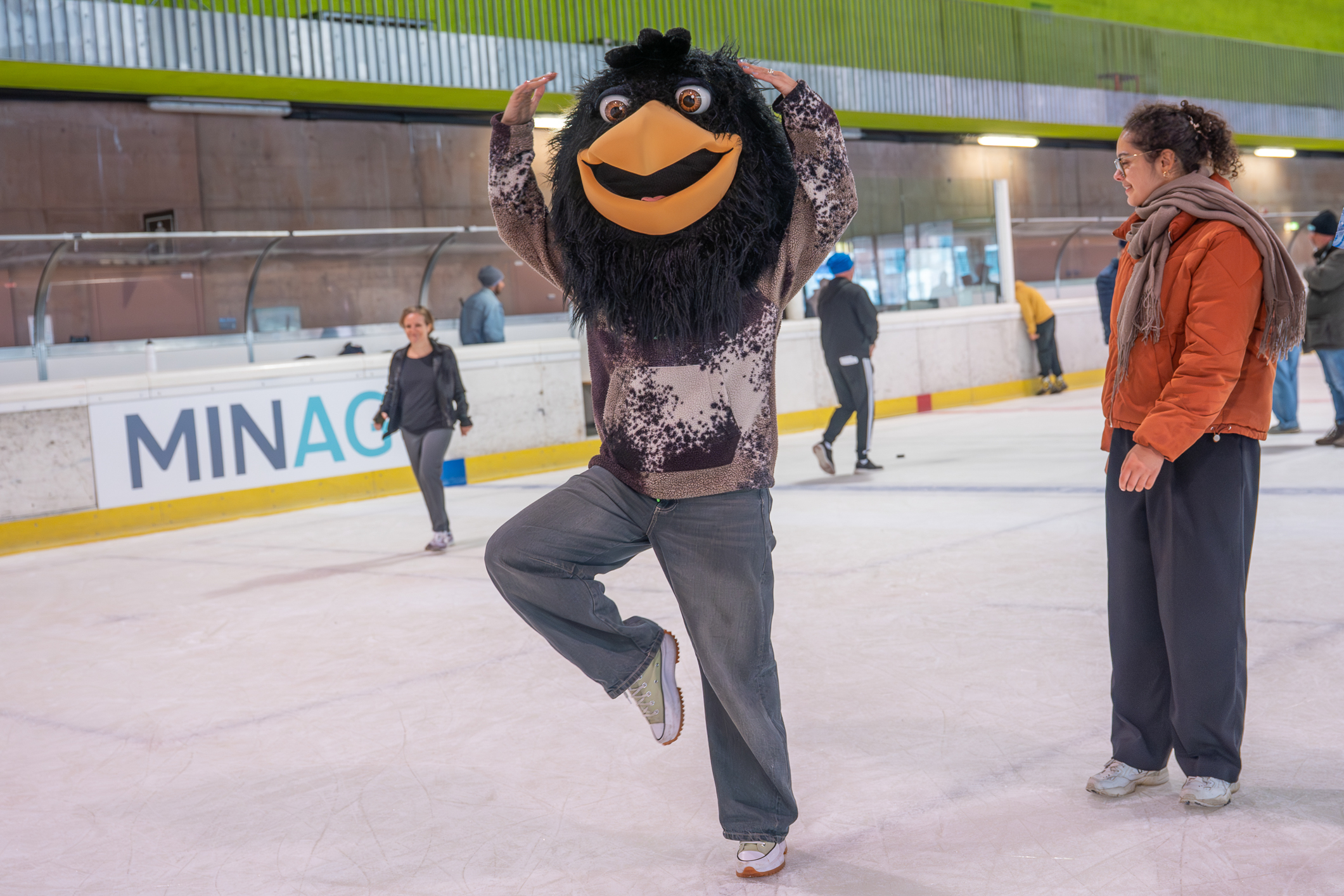 Person mit großem schwarzem Vogelkopf balanciert auf einem Bein auf dem Eis, daneben steht eine Frau in orangefarbener Jacke.
