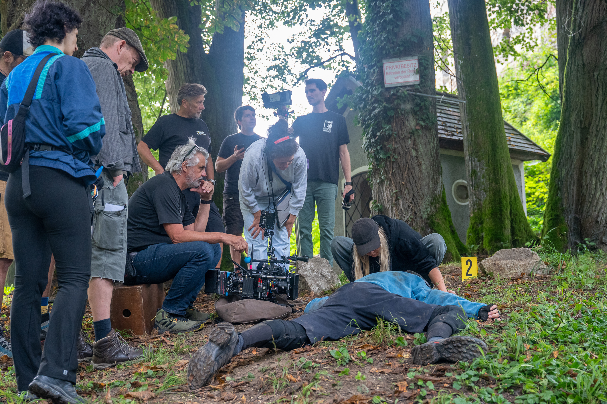 Ein Filmteam dreht im Wald eine Szene mit einer am Boden liegenden Person. Mehrere Crewmitglieder beobachten die Aufnahme.