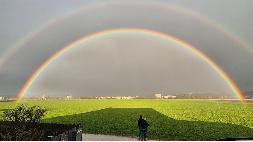 Doppelter Regenbogen über grünem Feld, im Vordergrund zwei Personen