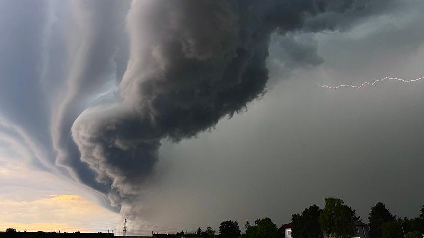 Beeindruckende Gewitterstimmung mit interessanten, dunklen Wolkenformationen, die sich von recht nach links auszubreiten scheinen. Rechts in der bereits dunklen Hälfte ist ein Blitz am Himmel.