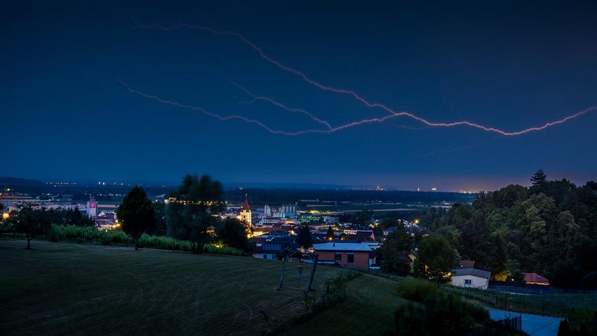 Eine Stadt von einer Anhöhe herab fotografiert. Einige Gebäude und Straßenlampen leuchten vereinzelt auf. Am dunklen Himmel zieht sich vertikal ein Blitz über die Landschaft.