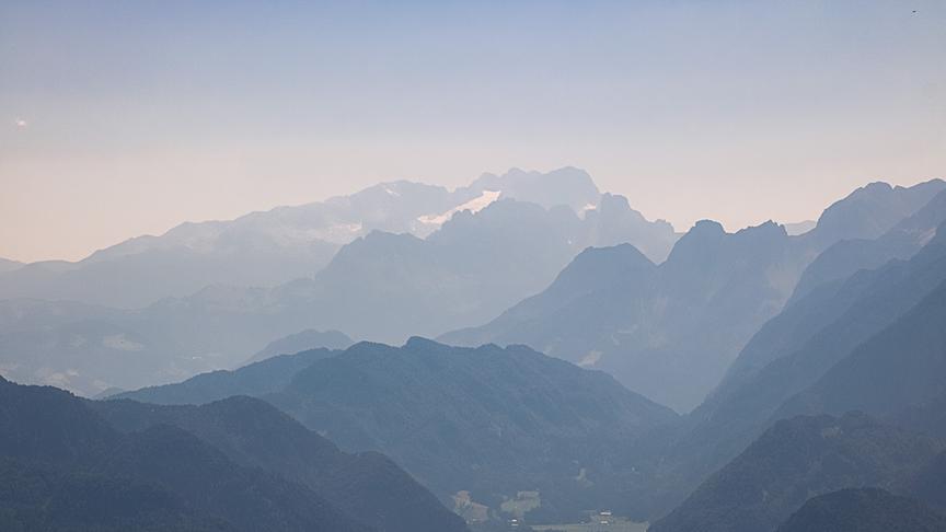 Bergkulisse mit hohen, zerklüfteten Bergen. Im Tal sind einige Häuser zu sehen mit viel Wald darum herum. Die Atmosphäre ist etwas getrübt und wirkt dunstig bei Schönwetterhimmel ohne Wolken.
