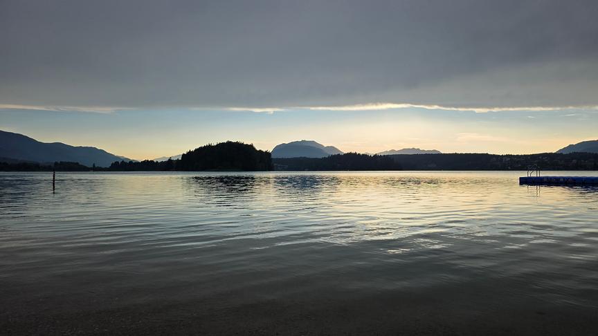 Weite Aufnahme eines Sees. Im Hintergrund erheben sich einige Hügel und Berge mit Wäldern empor. Über der Szene liegt eine geschlossene Wolkendecke im Vordergrund, während im Hintergrund noch blauer, hell erleuchteter Himmel ist.