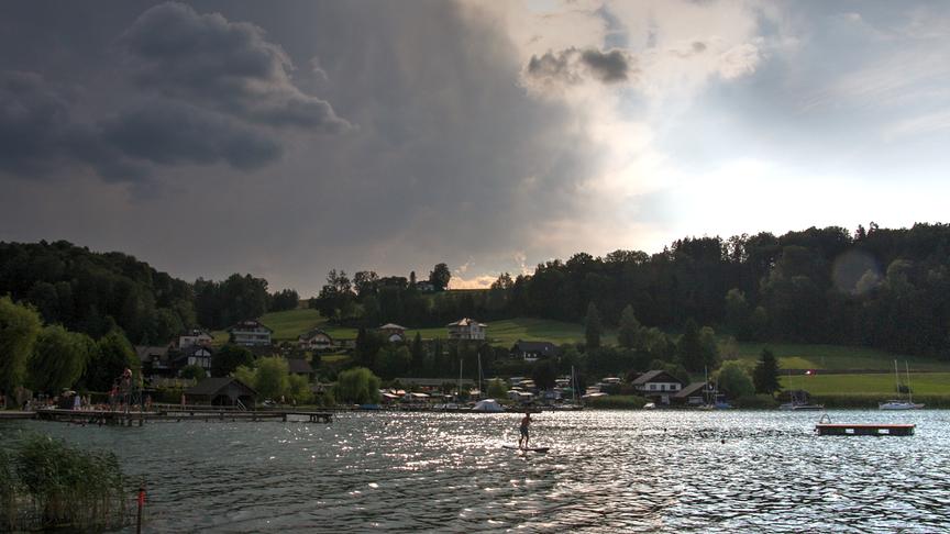 Ein See mit Badesteg und Menschen im Badekleidung. Ein Mann steht auf dem See auf einem Stand-up-Paddelbord. Im Hintergrund ist ein grün bewachsener Hügel mit Häusern. Von links zieht eine dunkle Wolkendecke auf, während rechts noch blauer Himmel ist.