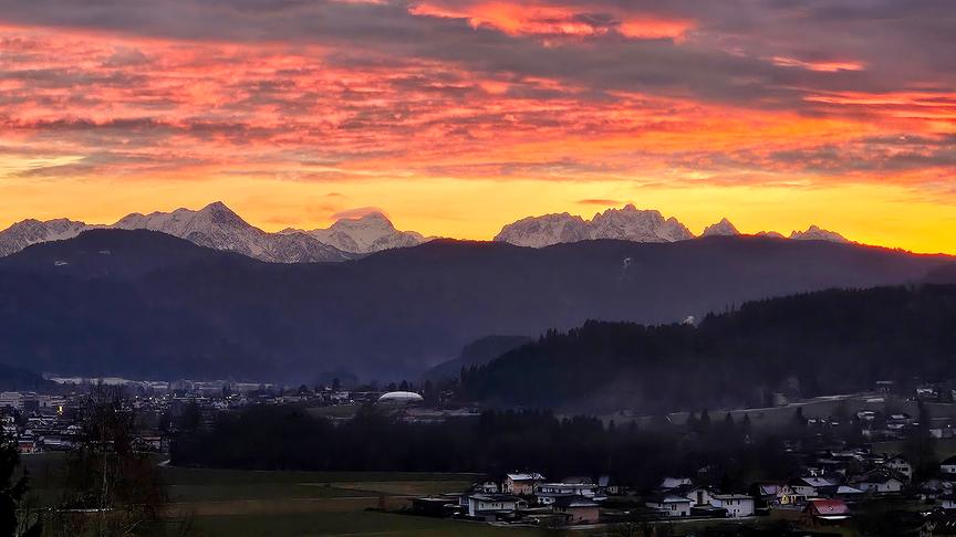 © Roswitha Groinig Kräftig rot-oranges Abendrot über einer Bergkette mit einer Siedlung im Vordergrund
