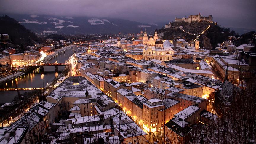 Die Stadtsalzburg liegt schneebedeckt und hell erleuchtet in der grauen Dämmerung.