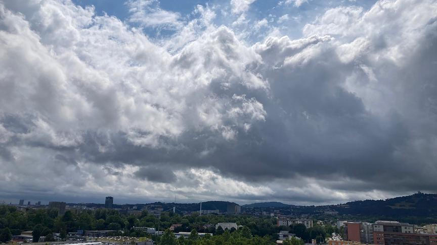 Ein weiter Blick über eine Stadtlandschaft unter einem dramatischen, wolkenverhangenen Himmel. Im Vordergrund erstreckt sich ein dichtes Band aus grünen Bäumen, aus dem vereinzelt Wohn- und Geschäftsgebäude sowie ein markanter dunkler Turm hervorstechen. Am rechten Bildrand erhebt sich im Hintergrund ein bewaldeter Hügel, auf dessen Spitze ein Gebäude zu erkennen ist. Der obere Teil des Bildes wird von einer massiven Wolkenfront dominiert, die von hellem Weiß bis zu tiefem Dunkelgrau reicht und nur stellenweise kleine Lücken zum blauen Himmel lässt.