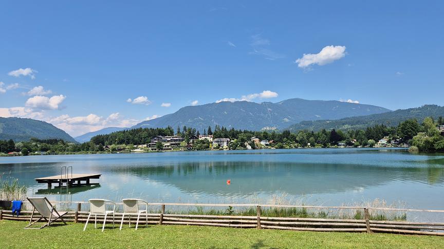 Ein idyllisches Landschaftsfoto zeigt einen ruhigen See unter einem strahlend blauen Sommerhimmel mit vereinzelten weißen Schönwetterwolken. Das klare, türkisfarbene Wasser spiegelt die bewaldeten Ufer und die dahinter aufragenden, sanften Berge wider.