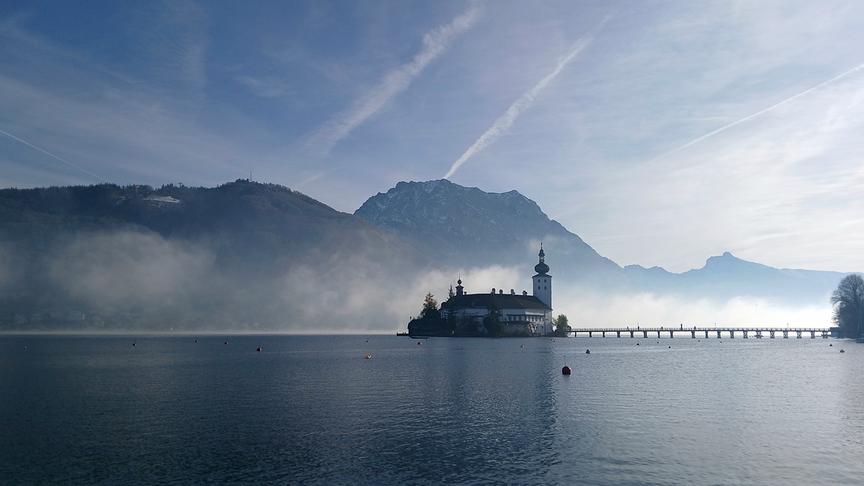 © Christa Katharina Dallinger Ein Schloss auf einer Insel in einem See hinter dem sich noch etwas Nebel befindet und im Hintergrund Berge erstrecken.