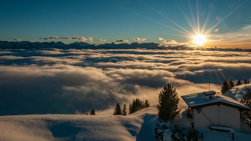 © Helmut Bischof Die Sonne scheint über eine dichte Wolkendecke hinweg auf einen verschneiten Berg