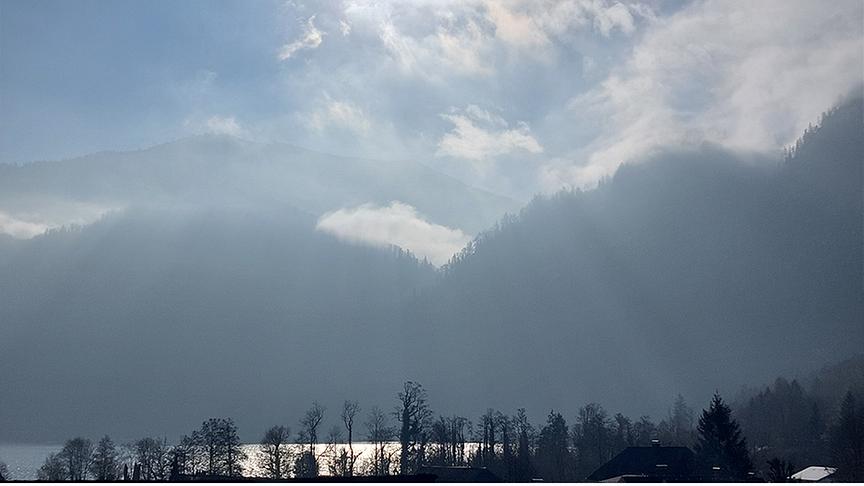 Durch die schleierartigen Wolken fallen sichtbare Sonnenstrahlen auf einen See.