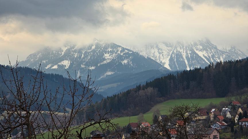 Im Tal ist es grün, auf den Bergen im Hintergrund liegt jedoch noch Schnee. Die Berggipfel hängen in grauen Wolken.