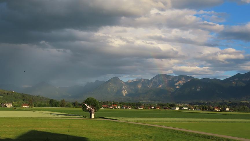 Gewitterwolken ziehen von links nach rechts über die Landschaft mit Wiesen und Bergen auf. Rechts noch ein Streifen blauen Himmels.