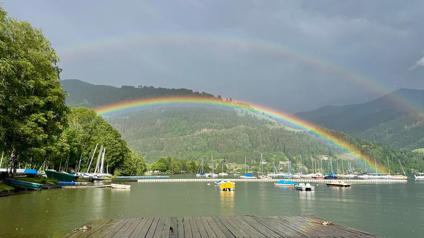 Zweifacher Regenbogen über einem See. Im Vordergrund erhellt die Sonne bereits, während der Himmel noch dunkel und grau wirkt.