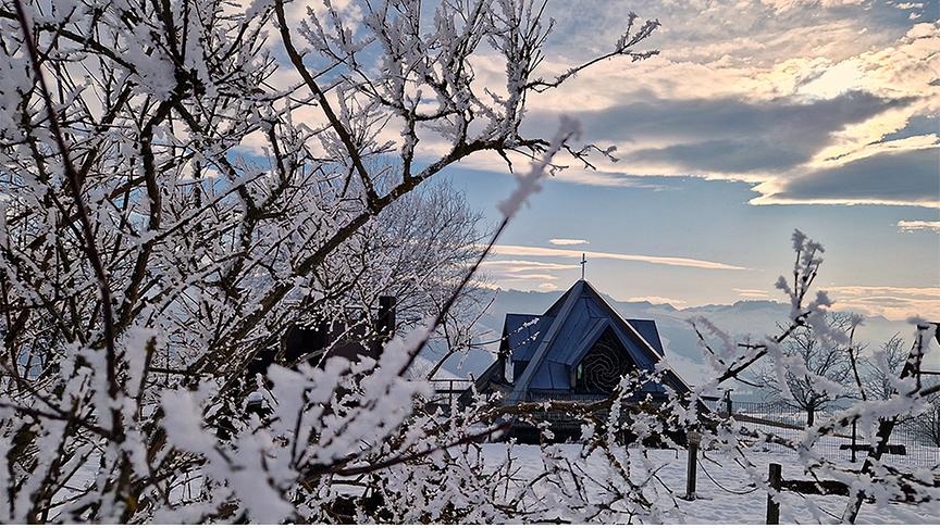 Durch schneebedeckte Ästchen hindurch sieht man eine Kirche mit Bergen im Hintergrund und Wolken am Himmel.