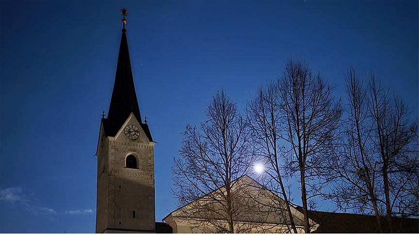 Heller Vollmond über einer Kirche, der durch die kahlen Bäume hindurch zu sehen ist