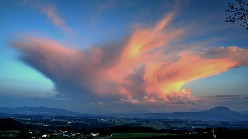 Hohe und ausladende Wolkenformation über einem Tal, die von der Sonne in organe-rot gefärbt wird.