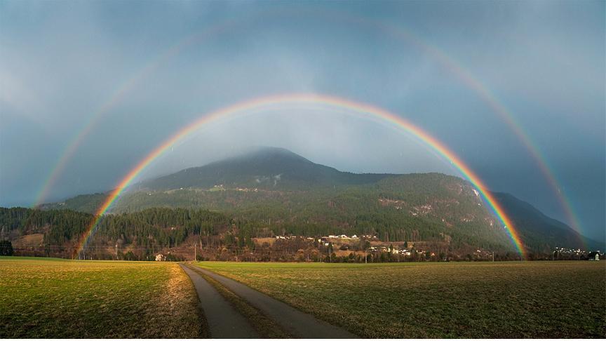 Doppelter Regenbogen über einem grünen Feld, im Hintergrund ein bewaldeter Berg
