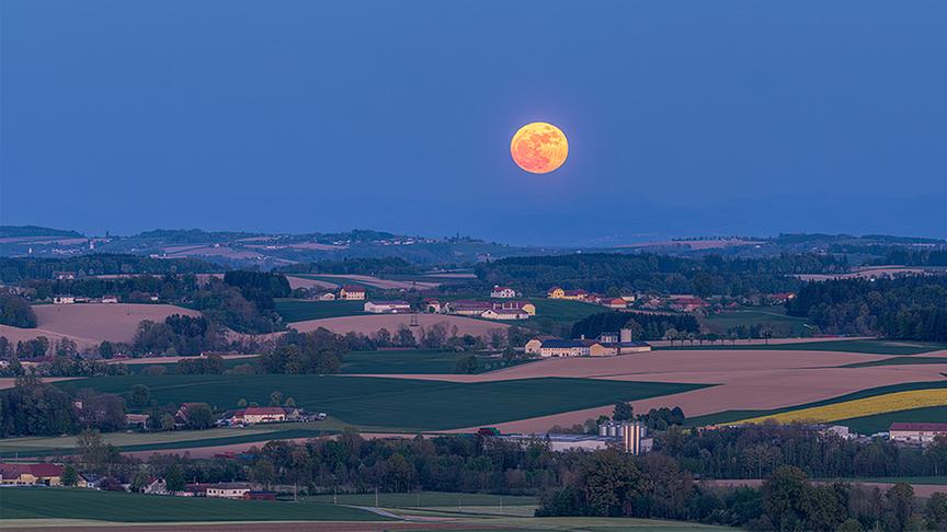 Gelb-oranger Vollmond steht über hügeliger Landschaft mit Feldern, Bäumen und vertstreuten Häusern.