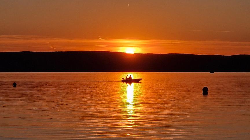 Der Sonnenuntergang färbt einen See in tiefes orange.Ein Boot mit zwei Personen schwimmt auf dem See, im Hintergrund ist die Silhouette einer flachen Hügelkette.